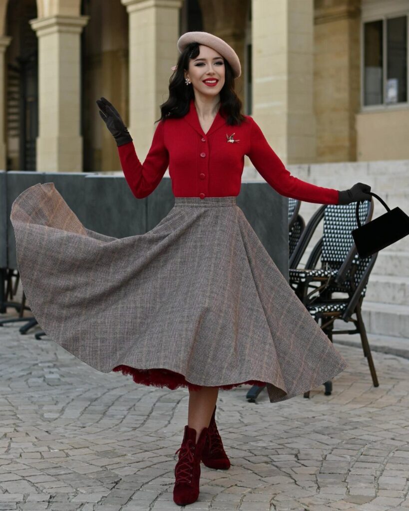 Woman in red cardigan and swirling brown plaid full skirt wearing a beret.