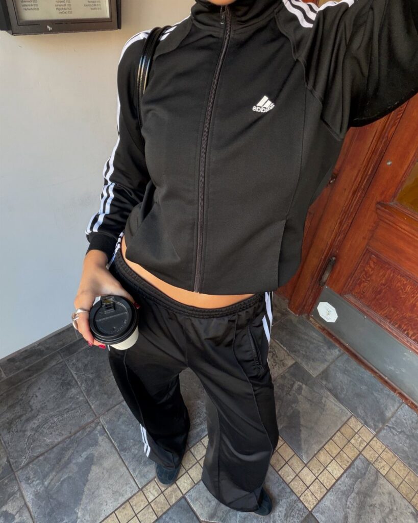 Person wearing a black Adidas tracksuit with white stripes and holding a coffee cup.