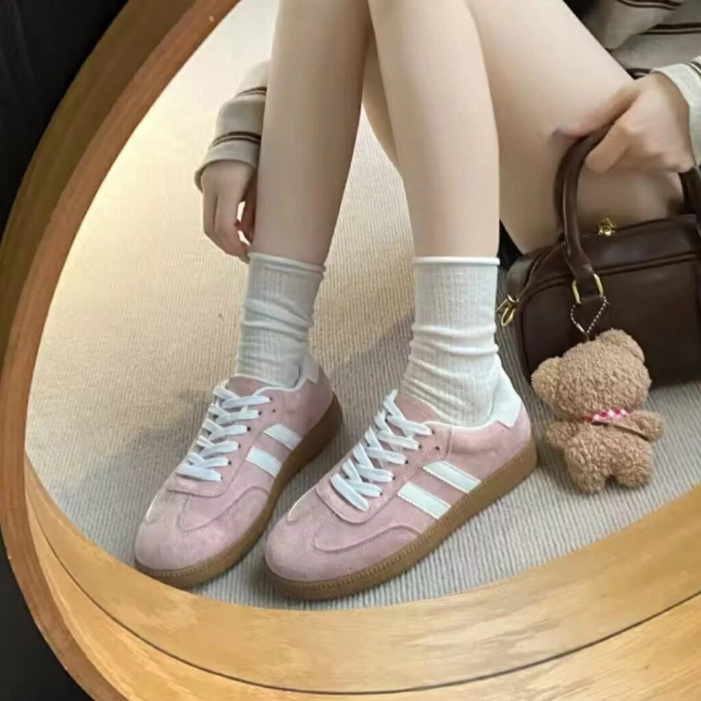 Pink suede tennis shoes with white stripes worn with white socks and a handbag.