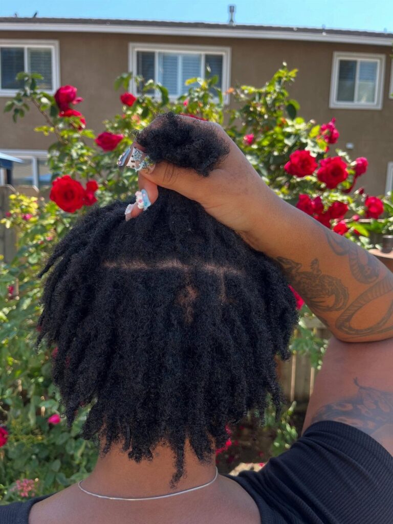 Person with short black locs posing in front of red rose bushes.