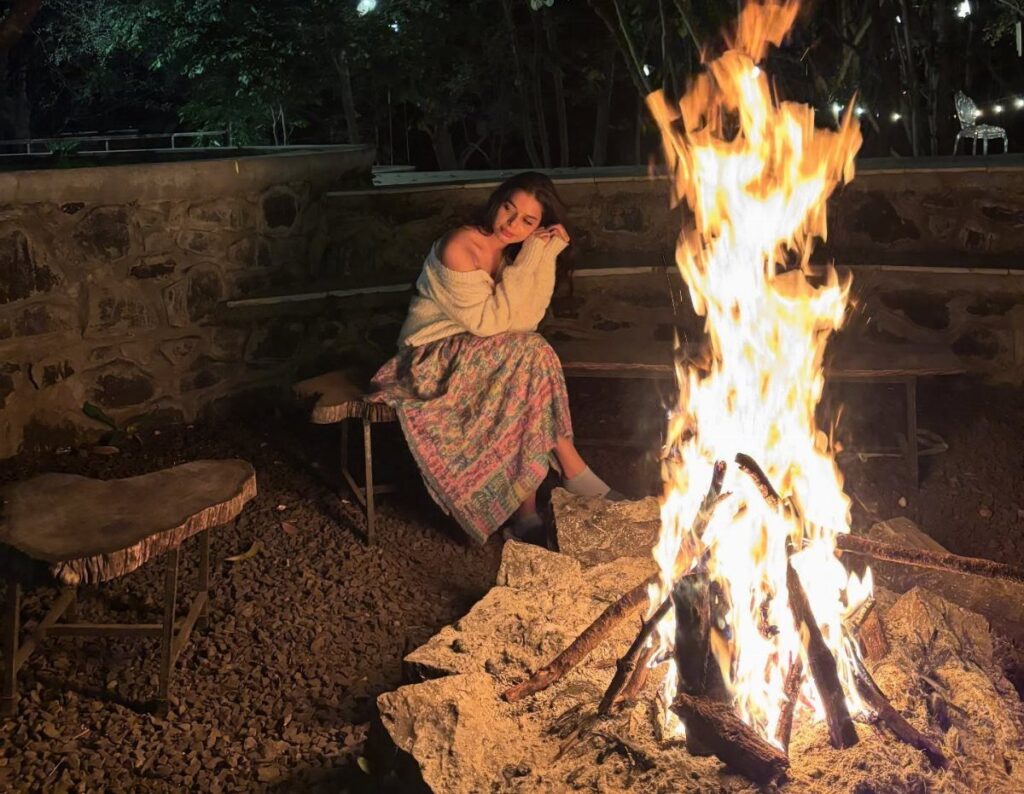 Woman in white sweater and colorful skirt sitting by a large outdoor bonfire