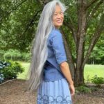 Woman with very long grey hair smiling outdoors in a blue denim outfit.