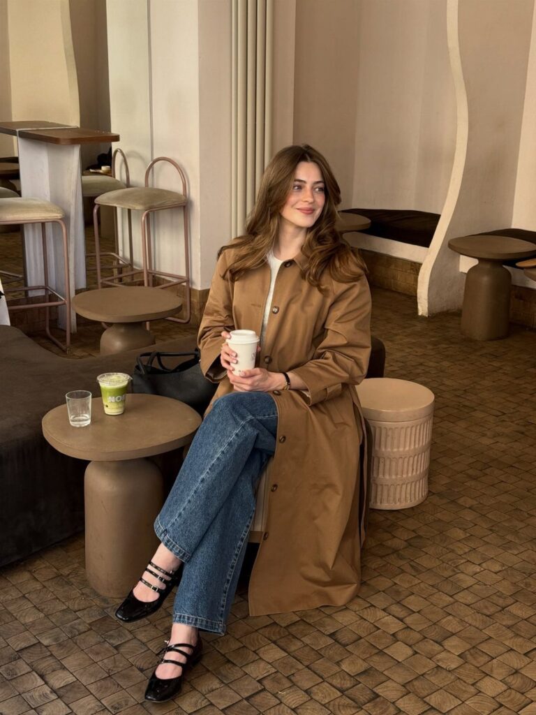 Woman in a tan trenchcoat and blue jeans sitting in a modern cafe.