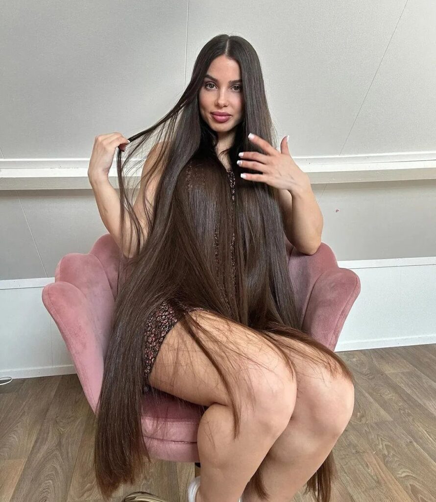 Woman with very long straight brown hair sitting in a pink velvet chair