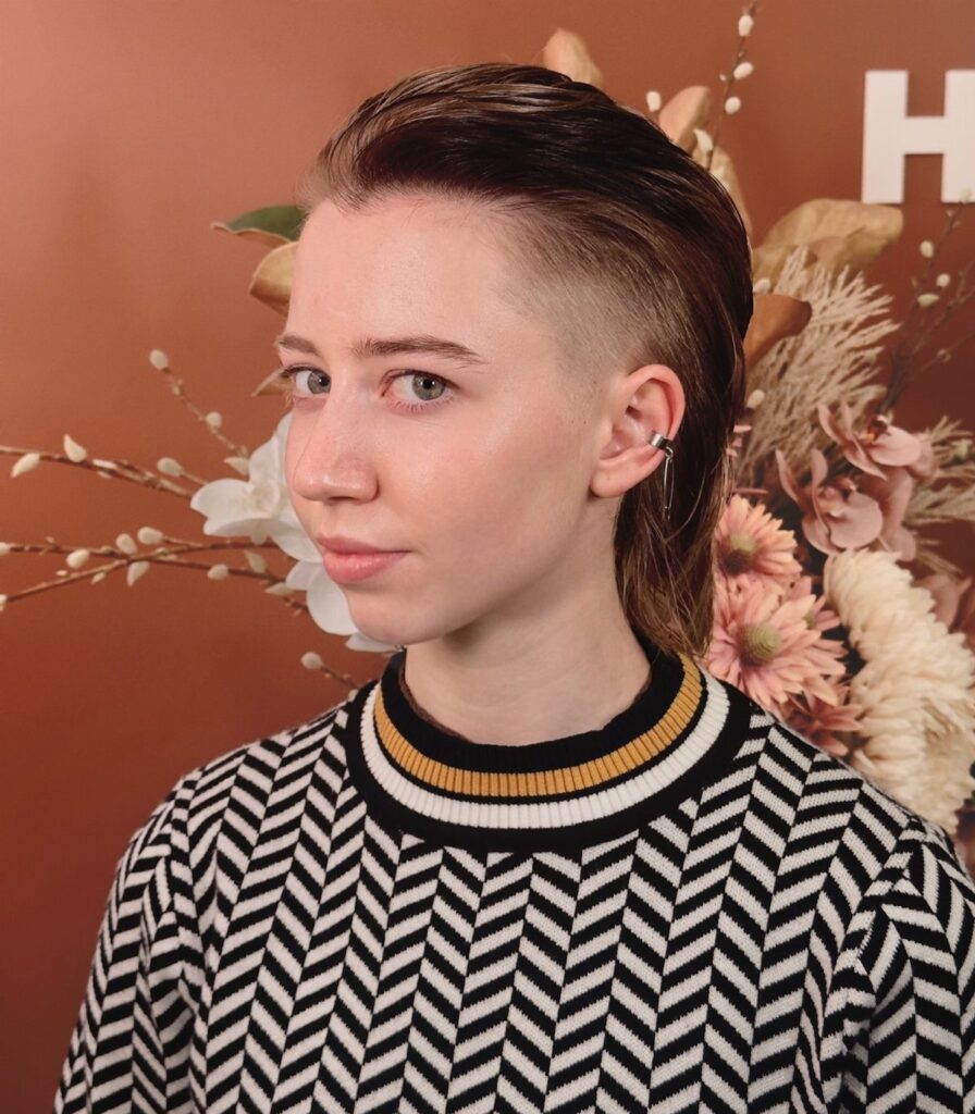 Person with a brown slicked-back textured mullet and shaved sides wearing chevron sweater.