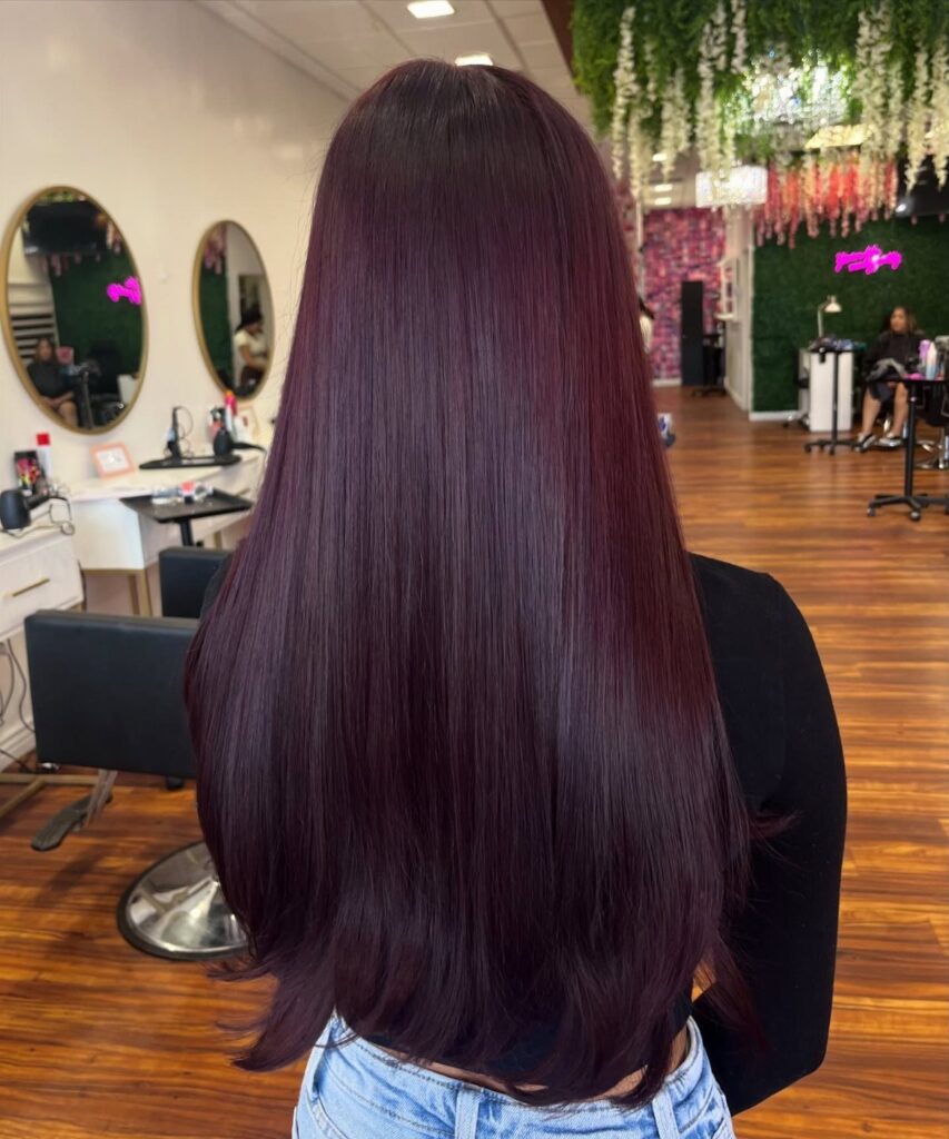 Long straight berry-purple hair shown from behind in a modern hair salon setting.