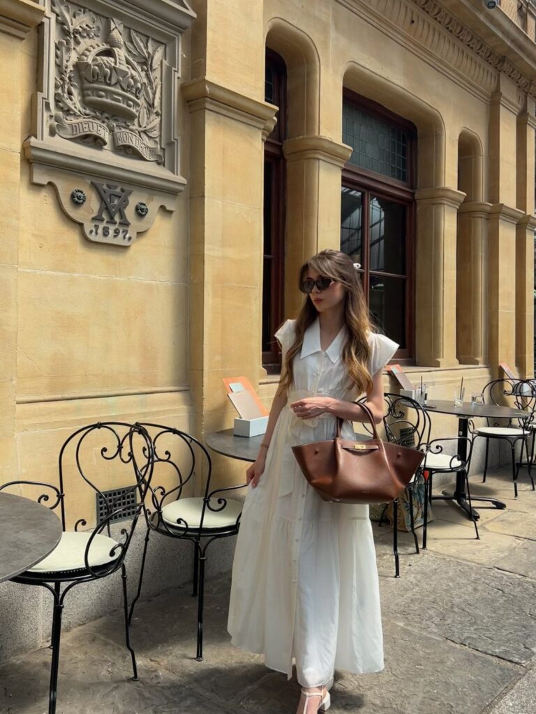 Woman in a long white cotton shirt dress standing by a stone building.