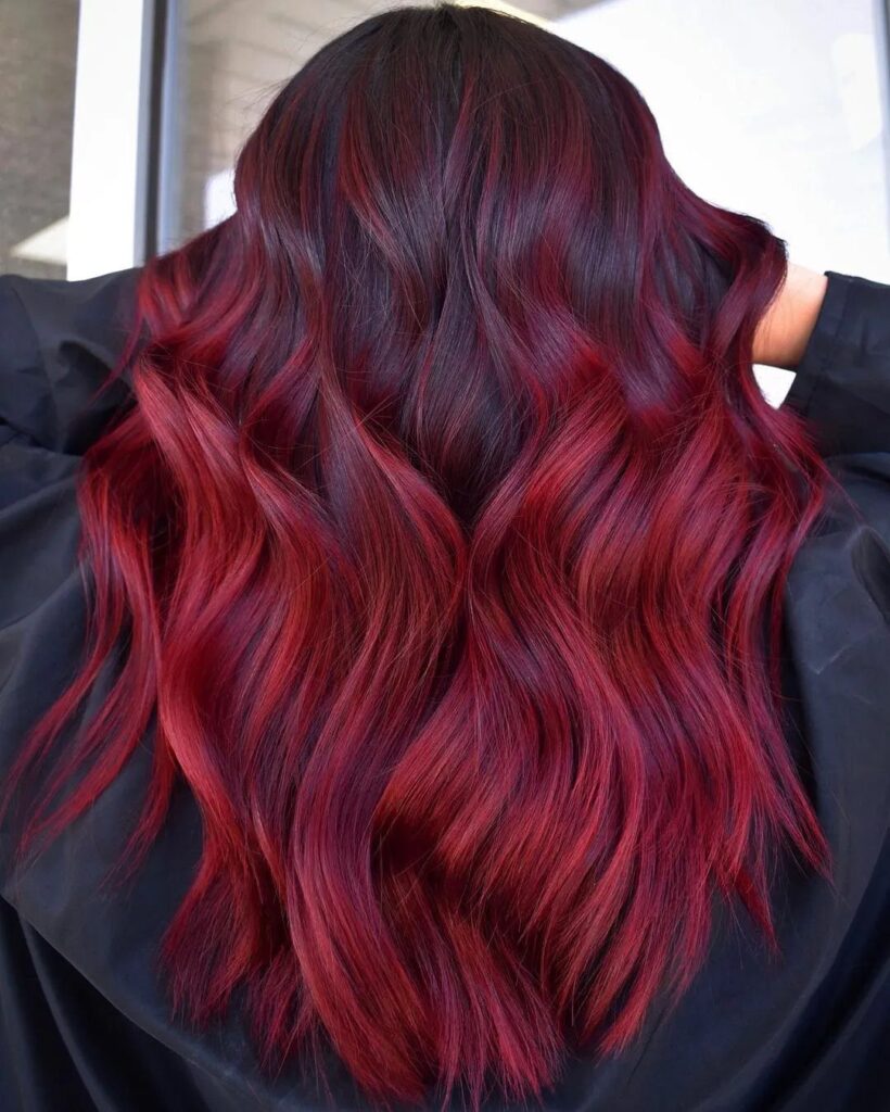 Long wavy hair with deep oxblood red roots transitioning to lighter red ends.